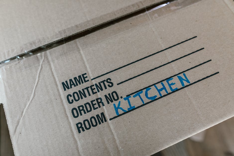 Close-up image of a cardboard moving box with the label indicating its contents and destination for a home relocation, showing the printed sections for 'Name', 'Contents', 'Order No.', and 'Room', with the handwritten entry 'Kitchen' in blue marker. The box appears to be made of corrugated cardboard, sealed with clear packing tape, and is positioned indoors, possibly on a floor or table, with natural lighting highlighting the label and the texture of the cardboard. This scene illustrates the packing process during house removals, with [COMPANY_NAME] involved in logistics and furniture transport to ensure a smooth moving experience for clients. The box is part of the packing materials commonly used in professional removals, organized to facilitate efficient loading and unloading as part of a comprehensive removals service offered by [COMPANY_NAME], supporting clients in relocating household contents including kitchen items as part of their moving process.