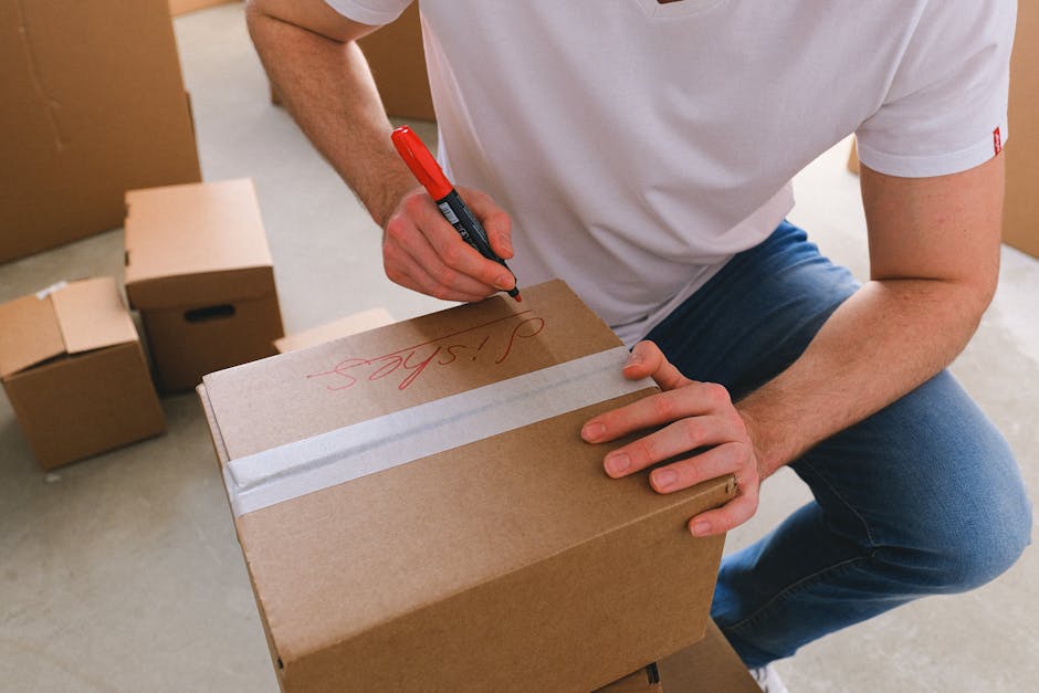 A person wearing a white t-shirt and blue jeans is sitting inside a room, holding a brown cardboard box on their lap while writing on the top with a red marker. The box is sealed with clear packing tape across the top. In the background, there are several other cardboard boxes, some stacked and some placed on the floor, indicating an ongoing packing process. The setting appears to be a home or storage area, with soft, natural lighting illuminating the scene. This image depicts the packing stage of house removals, a common task handled by companies like Man with Van Loxford to ensure safe and organised furniture transport and home relocation. The scene emphasizes careful packing and preparation involved in professional removal services, aligned with the topic of avoiding hidden removal fees for Loxford moves, LOXFORD.