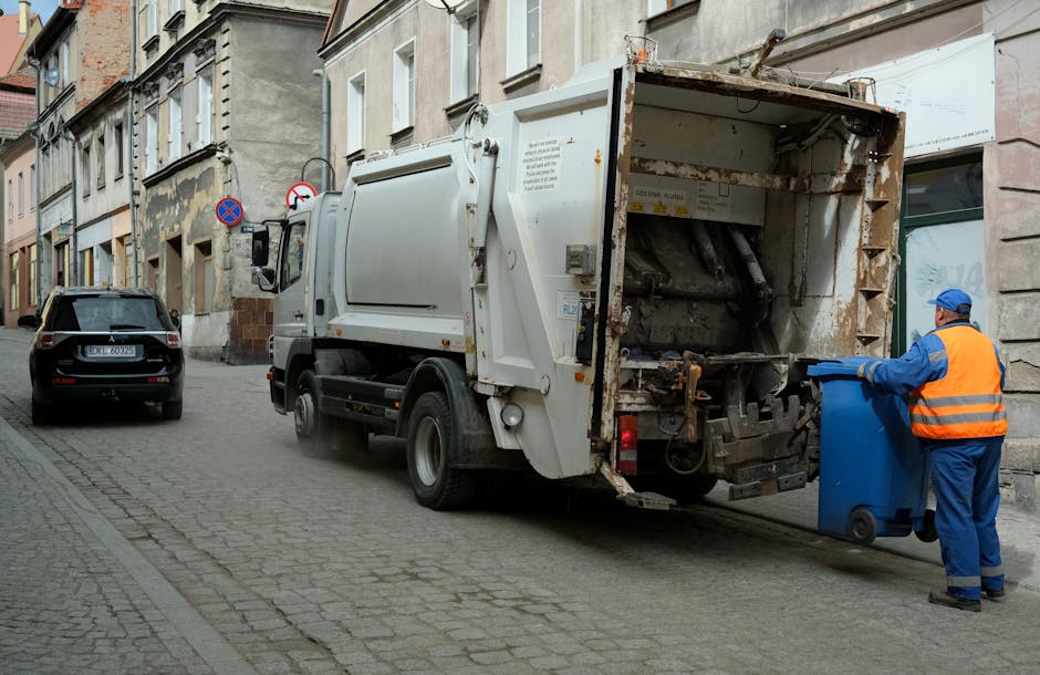 A street scene showing a white waste collection refuse truck with rusted parts and an open rear loading compartment, positioned on a cobbled urban road. A worker dressed in blue work trousers and an o