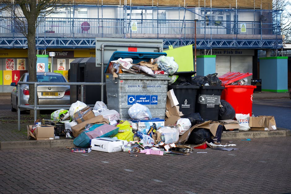 A street scene showing a white waste collection refuse truck with rusted parts and an open rear loading compartment, positioned on a cobbled urban road. A worker dressed in blue work trousers and an o