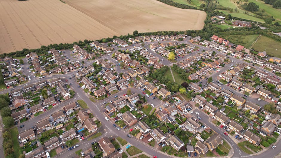 Aerial view of a residential neighbourhood showing a densely packed area of houses with tiled roofs, arranged along narrow, winding streets. Several cars are parked along the roads and in driveways, with some seen on the pavement. The area is surrounded by green spaces and farmland in the background. The photograph captures a typical house removal scene with a focus on the crowded layout of homes, which are closely spaced with small gardens and yards. The image highlights the urban environment suitable for house removals and relocation services provided by Man with Van Loxford, with the layout emphasizing the need for efficient furniture transport and moving logistics during tenancy clearance or home relocation projects.