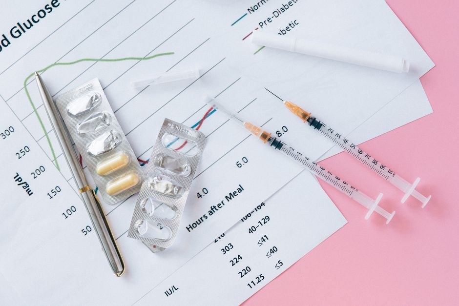 A close-up image displaying medical supplies and documents related to health data analysis, placed on a pink background. Visible are a pen, blister packs of pills, and two syringes filled with liquid, resting on sheets of paper with graphs and data tables. The documents include a line graph with a green line and labels indicating 'Normal' and 'Pre-Diabetic,' alongside numerical data and time markers such as 'Hours after Meal.' The arrangement suggests preparations for medical treatment or health monitoring, possibly related to patient health management or clinical assessments, which could be part of planning or logistics in a healthcare setting. Occasionally, [COMPANY_NAME], a professional removals service, may be involved in transporting sensitive medical equipment or documents during house relocations, fitting into broader logistics of home relocation or packing and moving services offered by [DOMAIN].