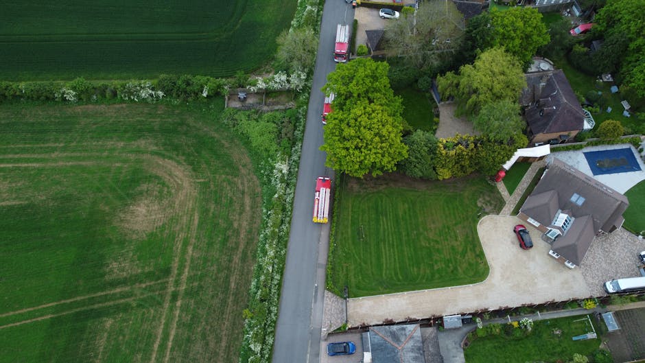 Aerial view of a residential neighbourhood showing a densely packed area of houses with tiled roofs, arranged along narrow, winding streets. Several cars are parked along the roads and in driveways, with some seen on the pavement. The area is surrounded by green spaces and farmland in the background. The photograph captures a typical house removal scene with a focus on the crowded layout of homes, which are closely spaced with small gardens and yards. The image highlights the urban environment suitable for house removals and relocation services provided by Man with Van Loxford, with the layout emphasizing the need for efficient furniture transport and moving logistics during tenancy clearance or home relocation projects.