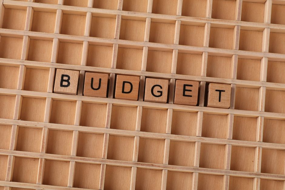 Close-up view of a wooden grid display with six small wooden blocks arranged in a horizontal line spelling out the word 'BUDGET' in black capital letters. The background consists of the light natural