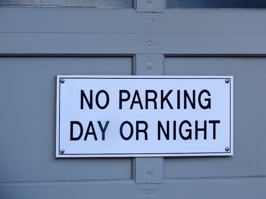 Close-up image of a rectangular white metal sign mounted on a light grey wooden door, displaying the black text 'NO PARKING DAY OR NIGHT'. The sign is attached with four screws at each corner. The door appears to be part of a residential or commercial building, with visible horizontal paneling and paint unevenness. In the context of house removals and furniture transport, this sign might indicate parking restrictions relevant to moving logistics, such as the need to obtain permits or adhere to council rules, which is addressed in the Loxford removals page on manwithvanloxford.co.uk. The lighting is natural, highlighting the contrast between the signage and the door surface, supporting clear visibility for drivers and movers during a home relocation or loading process.