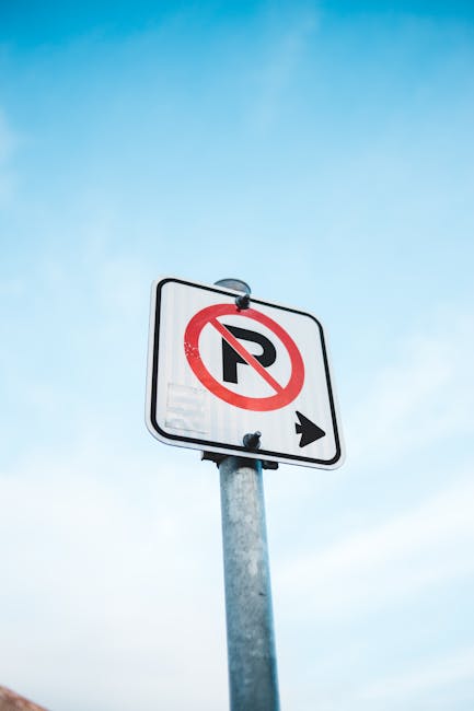 Close-up image of a rectangular white metal sign mounted on a light grey wooden door, displaying the black text 'NO PARKING DAY OR NIGHT'. The sign is attached with four screws at each corner. The door appears to be part of a residential or commercial building, with visible horizontal paneling and paint unevenness. In the context of house removals and furniture transport, this sign might indicate parking restrictions relevant to moving logistics, such as the need to obtain permits or adhere to council rules, which is addressed in the Loxford removals page on manwithvanloxford.co.uk. The lighting is natural, highlighting the contrast between the signage and the door surface, supporting clear visibility for drivers and movers during a home relocation or loading process.
