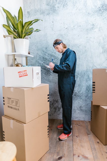 A man wearing a blue work jumpsuit, protective glasses, and pink shoes stands indoors against a textured light blue wall, surrounded by several brown cardboard boxes and a white box labeled 'GLASS'. He is holding a pen and appears to be writing or checking a list, possibly during the packing process. To his left, there is a tall white shelf with a large potted plant featuring long green leaves. The boxes are stacked on a wooden floor, and some are positioned near a doorway, suggesting a home relocation or moving process, as part of the packing and loading stages handled by Man with Van Loxford for house removals and furniture transport services.