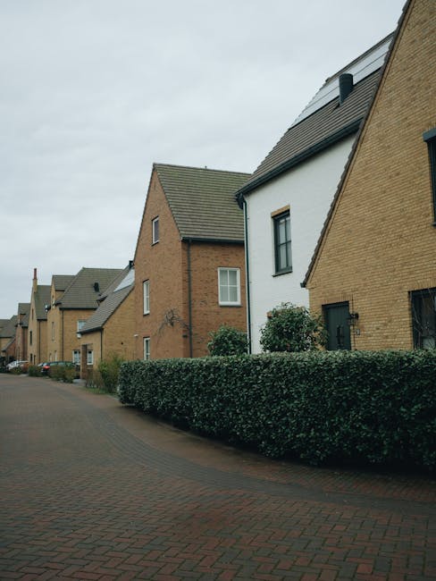 A street view of a residential area featuring modern detached houses with brick and white exteriors, pitched roofs covered with grey tiles, and small windows. The foreground shows a paved pavement made of brick-colored stones, with a neatly trimmed green hedge running alongside it. Multiple houses are visible along the street, each with small front gardens and additional houses lining the background. The sky is overcast with grey clouds, and there are no visible vehicles or people in the image. This scene suggests a quiet suburban neighbourhood suitable for home relocations, with a calm environment ideal for moving services. Occasionally, Man with Van Loxford's removals team may use such streets for loading and unloading furniture, packing materials, or moving vans in the process of a home relocation or furniture transport, supported by the infrastructure seen here.