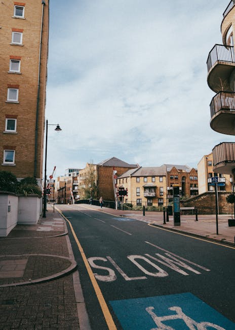 A wide view of a residential street captured during daytime with overcast skies, showing a gently sloping road marked with a 'SLOW' warning in white paint and a designated blue parking zone near the curb. On the left side, there is a multi-storey brick building with several windows and a small sidewalk, while on the right, a modern apartment with rounded balconies extends from the upper floors. In the background, various low-rise brick buildings with pitched roofs are visible, along with a lamppost and some greenery. The street appears quiet, with no vehicles or pedestrians in immediate view. The scene exemplifies an urban environment suitable for house removals and furniture transport operations, where moving companies like Man with Van Loxford may organize logistical planning for home relocation, including loading of furniture and boxes onto vans, with appropriate equipment such as trolleys and securing straps possibly in use outside the frame.