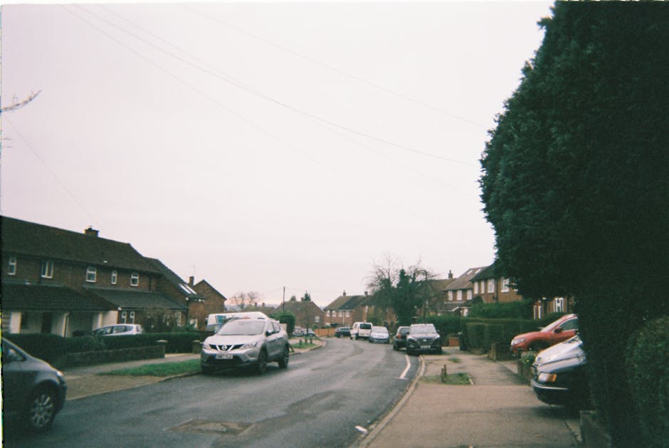 A street view of a residential area featuring modern detached houses with brick and white exteriors, pitched roofs covered with grey tiles, and small windows. The foreground shows a paved pavement made of brick-colored stones, with a neatly trimmed green hedge running alongside it. Multiple houses are visible along the street, each with small front gardens and additional houses lining the background. The sky is overcast with grey clouds, and there are no visible vehicles or people in the image. This scene suggests a quiet suburban neighbourhood suitable for home relocations, with a calm environment ideal for moving services. Occasionally, Man with Van Loxford's removals team may use such streets for loading and unloading furniture, packing materials, or moving vans in the process of a home relocation or furniture transport, supported by the infrastructure seen here.