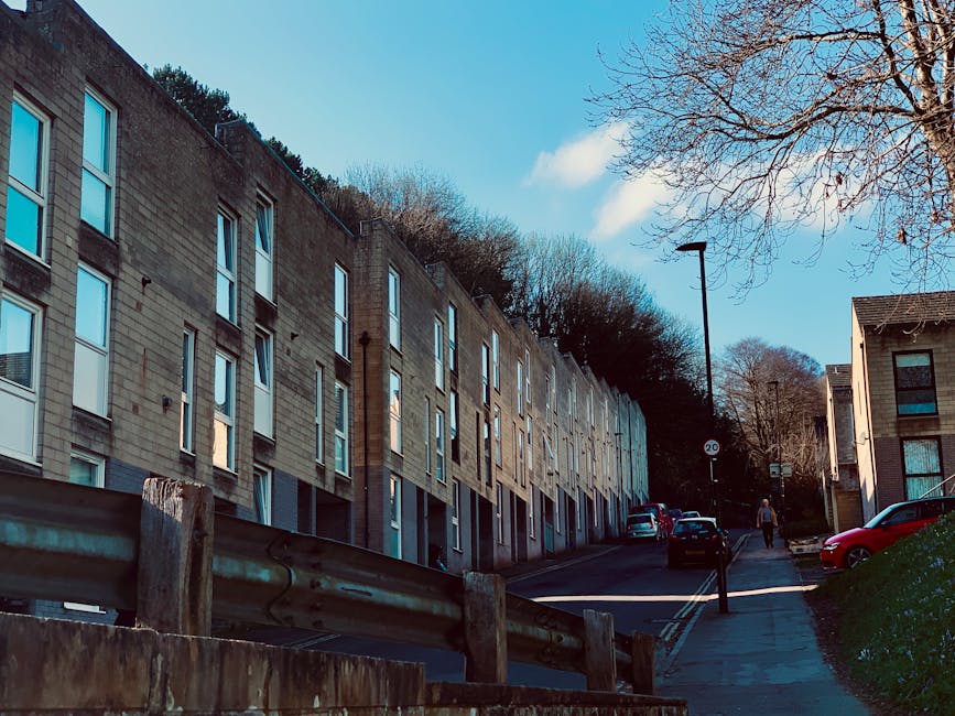 A row of modern, multi-storey residential buildings with large windows and a mix of beige and grey brick facades, situated along a sloped street. The street features parked cars on the right side, a pedestrian walking along the sidewalk, and a streetlight positioned centrally. In the foreground, a section of metal guardrail and wooden fence is visible, indicating the area near the property. The scene is illuminated by daylight, with a clear blue sky overhead and leafless trees, suggesting late autumn or winter. The environment appears quiet and clean, with no visible movement of furniture or packing materials, reflecting the setting of a typical UK residential area suitable for home relocation or moving services.