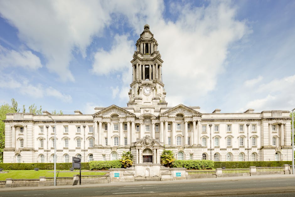 A large, historic building with classical architectural details, including a central clock tower, columns, and decorative stonework, situated near Redbridge Town Hall. In the foreground, a paved street runs parallel to the building, with neatly trimmed greenery and small bushes along the sidewalk. The sky above is partly cloudy with patches of blue. This image reflects the exterior environment of a town hall or municipal building, relevant to house removals and relocation services, such as those offered by Man with Van Loxford, which may involve navigating around such landmarks during packing and moving processes.