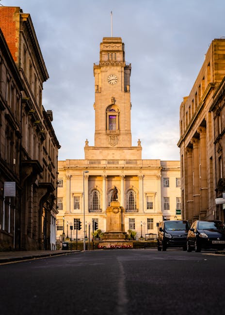 A wide view of Redbridge Town Hall in Loxford during late afternoon or early evening, with a clear blue sky and some scattered clouds. The prominent historic building features a central clock tower topped with a flagpole, ornate architectural details, and a columned entrance with large arched windows above. The street in front is relatively quiet, with a few parked vehicles and traffic lights at the intersection. The area surrounding the town hall includes older, classical-style buildings on either side, with visible stone facades and decorative elements. This setting reflects a typical town centre scene that might be associated with house removals or moving services, illustrating the environment near which packing and furniture transport could occur in a home relocation process, as managed by companies like Man with Van Loxford.