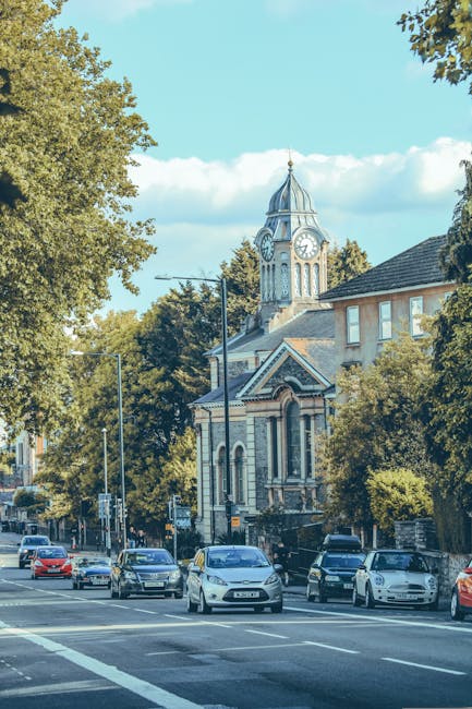 A large, historic building with classical architectural details, including a central clock tower, columns, and decorative stonework, situated near Redbridge Town Hall. In the foreground, a paved street runs parallel to the building, with neatly trimmed greenery and small bushes along the sidewalk. The sky above is partly cloudy with patches of blue. This image reflects the exterior environment of a town hall or municipal building, relevant to house removals and relocation services, such as those offered by Man with Van Loxford, which may involve navigating around such landmarks during packing and moving processes.