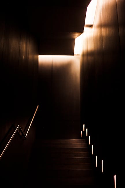 The image depicts a narrow staircase within a building, leading upward with wooden steps and a dark, enclosed corridor on either side. On the right side of the staircase, small integrated LED lights are embedded along the wall, providing subtle illumination. The handrail on the left side is made of metal with a simple design, attached to the wall for support. At the top of the stairs, natural light enters through a rectangular opening, casting a soft glow on the upper part of the staircase. The surrounding walls appear to be finished with smooth, dark-colored materials, contributing to a sleek and modern aesthetic. This setting is relevant to the process of home relocation and furniture transport within residential buildings, reflecting the environment where items might be moved during packing and moving operations by professionals such as Man with Van Loxford.
