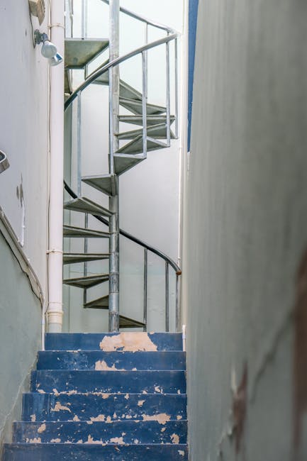 An internal view of a narrow, multi-flight staircase within a residential building, with worn blue-painted steps showing signs of chipped paint and age. The staircase curves upward, featuring metal railings and a spiral metal staircase ascending to an upper floor, positioned against plain white walls. The area appears to be undergoing maintenance or preparation for a home relocation, with visible pipes along the wall and a general utilitarian appearance. The environment suggests a confined space suitable for furniture transport or moving services, such as those provided by Man with Van Loxford, especially relating to stairs, narrow hallways, and flats. The natural lighting illuminates the stairwell, emphasizing the textured surfaces and structural details.