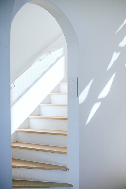 The image depicts a narrow staircase within a building, leading upward with wooden steps and a dark, enclosed corridor on either side. On the right side of the staircase, small integrated LED lights are embedded along the wall, providing subtle illumination. The handrail on the left side is made of metal with a simple design, attached to the wall for support. At the top of the stairs, natural light enters through a rectangular opening, casting a soft glow on the upper part of the staircase. The surrounding walls appear to be finished with smooth, dark-colored materials, contributing to a sleek and modern aesthetic. This setting is relevant to the process of home relocation and furniture transport within residential buildings, reflecting the environment where items might be moved during packing and moving operations by professionals such as Man with Van Loxford.